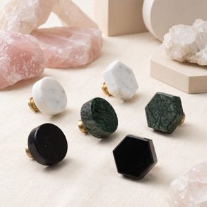 Puede incluir: Un conjunto de seis pomos para armarios con formas circulares y hexagonales. Los pomos están fabricados en mármol blanco, verde oscuro y negro, cada uno montado sobre una pequeña base de metal color latón, dispuestos sobre una superficie clara.