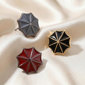 May include: Three umbrella-shaped stud earrings featuring smooth enamel finishes in dark gray, deep red, and glossy black. Each piece has a metallic frame that defines the eight panels of the umbrella canopy, with a central point and post back.