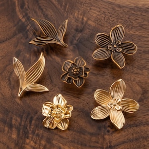 May include: A collection of six metal cabinet knobs featuring floral and leaf designs in varying shades of gold and bronze. The set includes two leaf-shaped knobs, two five-petaled flower knobs, and two multi-layered flower knobs with textured details.