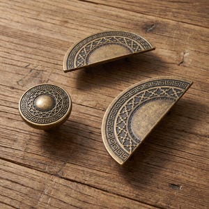 May include: A set of three antique brass-toned cabinet hardware pieces, including one circular knob and two semi-circular drawer pulls. Each piece features a raised Greek key border pattern and a central intricate lattice design in dark metal.