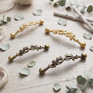 May include: Four metal cabinet pulls with a vine and leaf design. Two pulls feature a bright gold finish, while the other two have a darker, antique bronze finish. Each pull has a curved center section with detailed leaves and spherical ends.