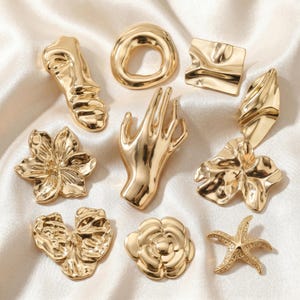 May include: A collection of ten gold-toned metal charms featuring various shapes including a human hand, a face, a starfish, a flower, a camellia, and abstract organic forms with a polished, reflective surface and smooth, flowing contours.