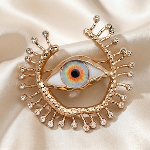 Può includere: Una spilla in metallo dorato a forma di occhio con sclera bianca e iride multicolore nei toni dell'arancione, giallo e blu. La montatura presenta una texture martellata ed è decorata con numerosi piccoli strass trasparenti a taglio rotondo.