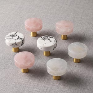 Peut inclure: Un ensemble de sept boutons de placard circulaires avec des dessus en pierre polie dans des tons rose pâle, blanc avec des veines noires et blanc translucide, chacun monté sur une petite base cylindrique en métal doré brossé.