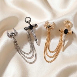May include: Two lapel pins featuring a winged skull and a sword with a black centre stone and clear rhinestones. One pin is silver-toned with silver chains, and the other is gold-toned with gold chains, both connected by three draped metal chains.