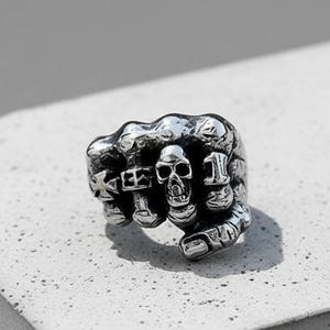 May include: A silver-colored ring shaped like a clenched fist, featuring a skull on the knuckles. The ring has the word "KEEP" and the number "1" engraved on it. The ring is set against a gray background.