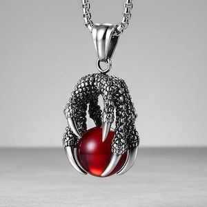 May include: A silver-colored pendant necklace featuring a claw design grasping a red orb. The claw has intricate detailing, and the chain is a classic link style. The pendant hangs against a neutral background, highlighting the jewelry's design.