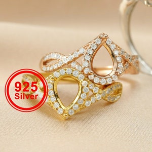 May include: Three silver rings with a teardrop-shaped setting for a gemstone. The rings are adorned with small, clear stones. The ring on the left is gold, the middle ring is rose gold, and the ring on the right is silver. The text "925 Silver" is visible on the left side of the image.