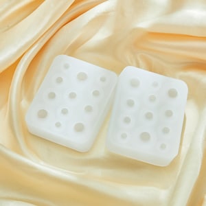 以下が含まれることがあります： さまざまなサイズの円形インデントが付いた、2つの白い長方形のシリコン型。 金色のサテン生地の上に型が配置されています。 この型は、おそらく工芸品や樹脂アート作品の作成に使用されます。