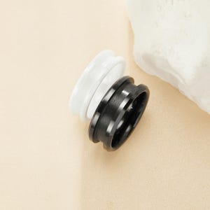 May include: Two rings, one black and one white, are stacked on top of each other. The rings have a cylindrical shape with a grooved design. The background is a light beige color with a white rock.