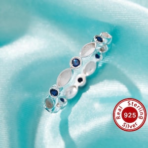 May include: A silver ring with a row of blue gemstones set in a delicate, oval-shaped design. The ring is on a light blue background with the words "Real 925 Sterling Silver" in a red circle.