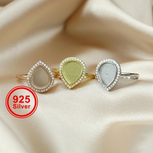 Op de afbeelding: Drie traanvormige ringen met heldere stenen in een pavé-setting. De ringen zijn gemaakt van zilver, goud en roségoud. De ringen liggen op een lichtbruine stoffen achtergrond. 925 Zilver.