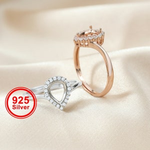 May include: Two silver rings with teardrop-shaped settings, one with a clear stone and the other with a clear stone setting. The rings are on a light beige fabric background. The text "925 Silver" is in a red circle.