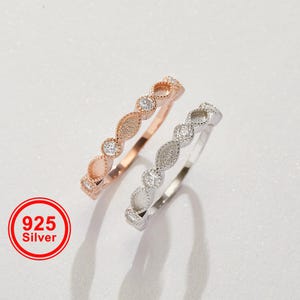 Puede incluir: Dos anillos de plata, uno de oro rosa y otro plateado, con un patrón repetido de piedras claras redondas y en forma de marquesa. Los anillos están etiquetados como "925 Silver" en un círculo rojo.