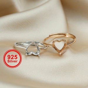May include: Two silver rings with heart-shaped settings. The ring on the left is silver, and the ring on the right is gold. The rings are on a beige fabric background. 925 Silver.