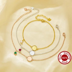 May include: Three silver chain bracelets with round, white, and gold charms. The charms have a textured surface. The bracelets are layered on top of each other. The bottom bracelet has a red gemstone charm. The top bracelet has a green gemstone charm. The middle bracelet has a gold charm. The image includes the text "Real Sterling 925 Silver".