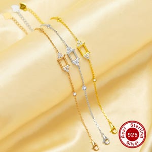Könnte beinhalten: Drei silberne Armbänder mit einem quadratischen Anhänger, der mit kleinen Zirkonia-Steinen besetzt ist. Die Anhänger sind in verschiedenen Größen und an einer zarten Kette befestigt. Die Armbänder liegen auf einem goldenen Untergrund.