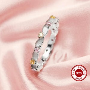May include: A silver ring with a repeating pattern of marquise-shaped silver elements and alternating pink and yellow gemstones. The ring is set against a pink background and features a "Real Sterling 925 Silver" stamp.