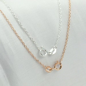 May include: Two delicate chains, one silver and one rose gold, lie on a white surface. Both chains feature a lobster clasp closure.