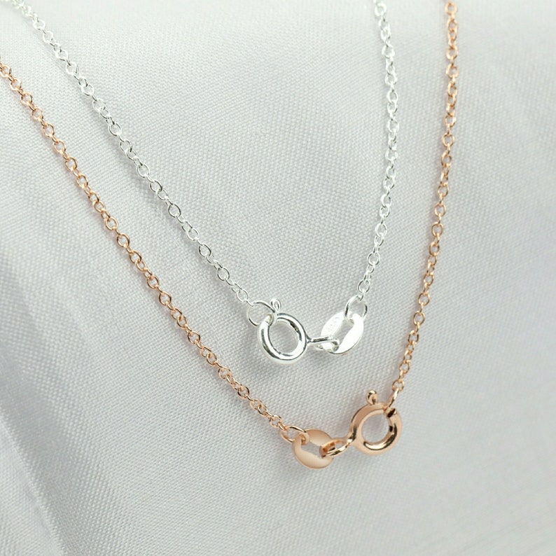 May include: Two delicate chains, one silver and one rose gold, lie on a white surface. Both chains feature a lobster clasp closure.