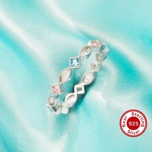 May include: A silver ring with a repeating pattern of alternating shapes and colored gemstones. The ring features square, oval, and diamond-shaped elements, with pink, blue, and clear stones. The ring is set against a light blue satin background.