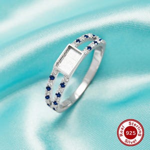 May include: A silver ring with a square bezel set with blue stones. The ring is made of sterling silver and has a double band design. The ring is on a blue background.