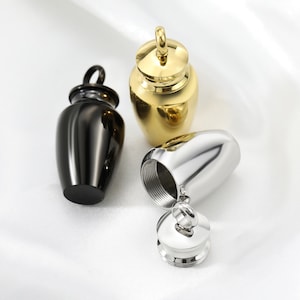 Puede incluir: Tres pequeñas urnas de metal con tapas. Una urna es dorada, una es negra y una es plateada. La urna plateada está abierta, revelando el interior.