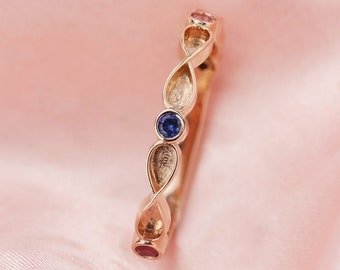 Puede incluir: Un anillo de banda de oro con un diseño delicado y retorcido. El anillo presenta tres pequeñas piedras preciosas: un zafiro azul y dos zafiros rosas.