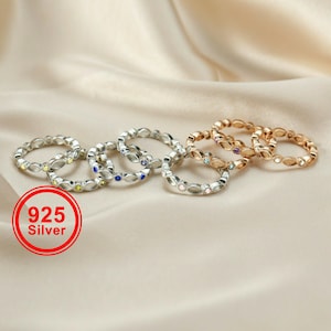 May include: A collection of silver and gold-toned rings with small, colorful gemstones. The rings are arranged on a cream-colored fabric. The image includes a red circle with the text "925 Silver".