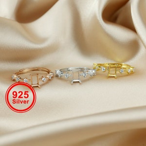 Pode incluir: Três anéis de prata com uma configuração quadrada para uma pedra preciosa. Cada anel tem uma fileira de pequenas pedras transparentes ao redor da configuração. Os anéis estão em um fundo de tecido marrom claro. O texto "925 Silver" está em vermelho em um círculo branco.