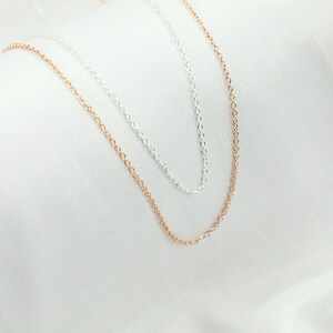 May include: Two delicate chain necklaces, one silver and one rose gold, lie on a white fabric background.