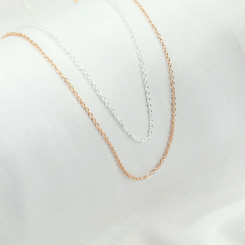 May include: Two delicate chain necklaces, one silver and one rose gold, lie on a white fabric background.