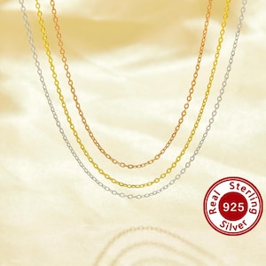 May include: Three silver chains, one gold chain, and one rose gold chain layered on a cream-colored background. The chains are all the same style, with small, oval links. A red circle with the text "Real 925 Sterling Silver" is in the lower right corner.