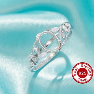 Puede incluir: Un anillo de plata de ley con un engaste ovalado, adornado con detalles de hojas y marquesas. El anillo se presenta sobre un fondo de satén azul claro, con un sello rojo y blanco "Real Sterling 925 Silver".
