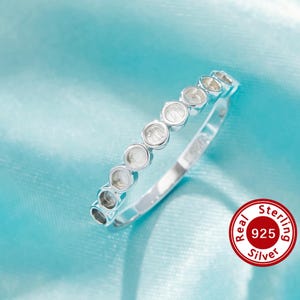 Pode incluir: Um anel de prata com uma fileira de engastes circulares, possivelmente para pedras preciosas, sobre um fundo azul claro. O anel é marcado com um selo "Real Sterling 925 Silver".