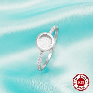 May include: A silver ring with a round, textured centre and a band of small clear stones. The ring is on a turquoise background. Real Sterling 925 Silver.