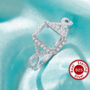 May include: A silver ring with a square-shaped setting for a gemstone. The ring is adorned with small, clear stones and has a twisted band.