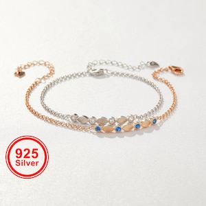 May include: Two delicate chain bracelets, one silver-toned and one rose gold-toned, featuring marquise-shaped accents. The silver bracelet has clear stones, while the rose gold bracelet features blue stones. A red circular logo reads 925 Silver.