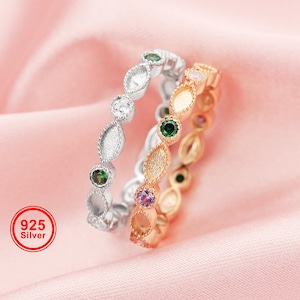 May include: Three silver and gold rings with gemstone accents. The rings feature a repeating pattern of oval and round settings, each holding a different colored gemstone. The rings are set against a pink background, with a "925 Silver" stamp.