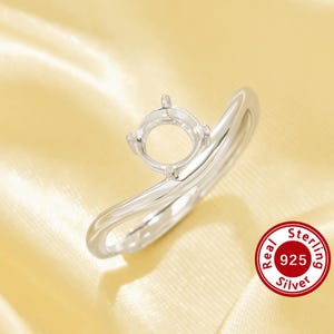 May include: A silver ring setting featuring a four-prong mount for a gemstone. The band has a smooth, polished finish and a curved, wave-like design. A circular red stamp in the corner displays the text Real Sterling 925 Silver.