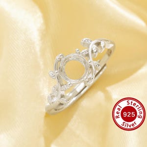 May include: A silver ring setting featuring a circular centre mount for a gemstone, surrounded by a delicate vine and leaf design along the band. The metal is marked as 925 sterling silver, indicating the composition of this jewellery component.