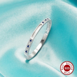 May include: A silver band ring with a row of small blue gemstones. The ring is on a light blue background. The text "Real Sterling 925 Silver" is visible in the lower right corner of the image.
