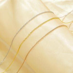May include: Three gold chains of different colors, silver, gold, and rose gold, are laid out on a cream-colored fabric background.