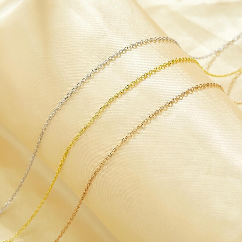 May include: Three gold chains of different colors, silver, gold, and rose gold, are laid out on a cream-colored fabric background.