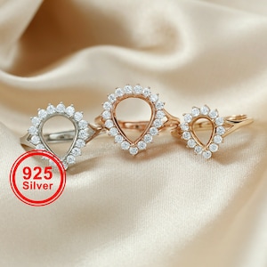May include: Three silver rings with a teardrop-shaped opening in the centre, surrounded by small clear stones. The rings are on a beige fabric background. 925 Silver.