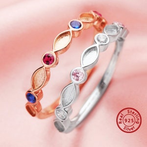 May include: Two sterling silver rings with colorful gemstones. The top ring is rose gold with red, blue, and clear gemstones. The bottom ring is silver with pink and clear gemstones. The rings are on a pink background.