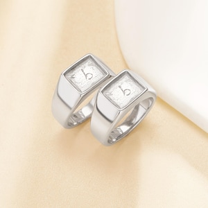 Puede incluir: Dos anillos de sello plateados con una cara cuadrada. Cada anillo presenta la letra "b" en un cuadrado claro y texturizado. Los anillos se exhiben sobre una superficie beige claro, mostrando sus superficies pulidas y reflectantes. Los anillos son un artículo de joyería clásico.