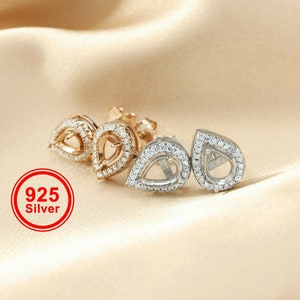 May include: A pair of silver teardrop-shaped earring settings with a clear stone setting. The earrings are surrounded by small clear stones. The earrings are on a light brown fabric background. 925 Silver.