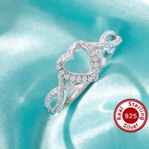 May include: A silver ring with a heart-shaped design and a row of small clear stones around the heart. The ring is on a light blue background. Real 925 Sterling Silver.