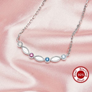 May include: A silver necklace with a delicate chain and five oval-shaped pendants. Each pendant features a small, clear gemstone in the center, with a white opal at the top and a pink, blue, and light blue gemstone in the other pendants. The necklace has a "Real Sterling 925 Silver" tag.